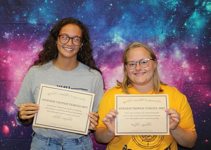 two students hold up certificates in front of a cosmic space background